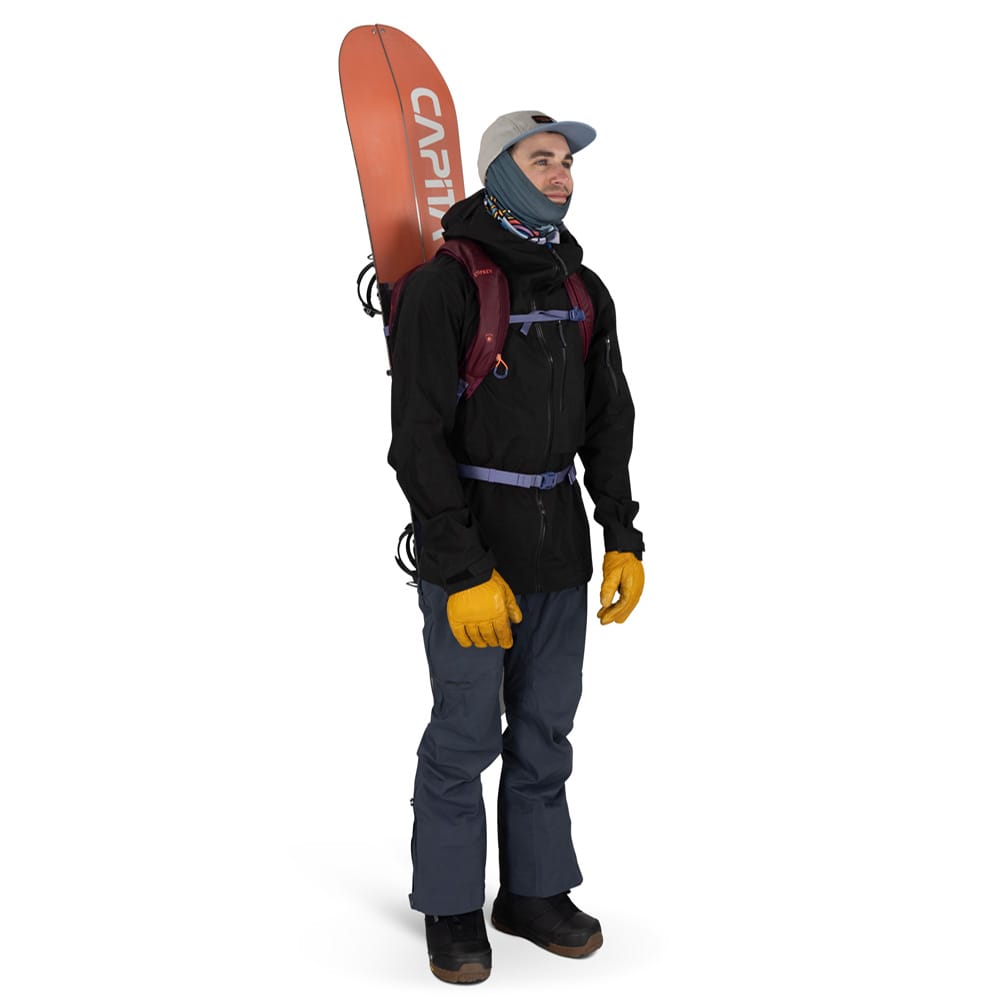 en mann bærer en Osprey Glade 12 ski-sekk (unisex) med et snowboard